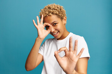 Wall Mural - Attractive black cheerful girl with blonde hair having fun on blue background doing ok sign with hand on eye looking through fingers, dressed in white mockup t-shirt. Body language, symbols, gestures