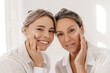 © Look! - Close-up of two attractive young women looking at camera applying cream to their face on white background. Blonde ladies in casual wear. Beauty needs care concept