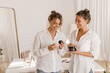 © Look! - Happy young caucasian women holding decorative cosmetics for testing indoors. Blondes wear white shirts and jeans rejoice in acquisition. Home spa concept