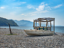 On The Shore There Is A Shed And An Old, White, Wooden Boat. View Of The Mountains, The Sea. Blue Sky. Seascape. On The Shore There Are Large Stone Pebbles. Desktop Wallpaper. Copy Space. Horizontally