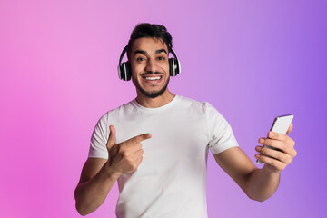 Poster - Portrait of funky smiling young Arab man in wireless headphones pointing at smartphone in neon light