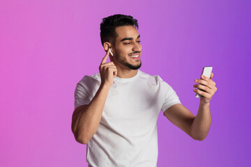 Wall Mural - Cool young Arab man listening to music in wireless earphones, looking at smartphone screen in neon light