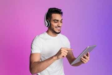 Poster - Positive young Arab man having video call, using tablet pc, wearing wireless headphones in neon light