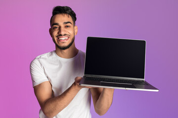 Wall Mural - Smiling millennial Arab man showing laptop computer with empty black screen in neon light, mockup for website or ad