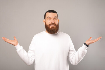 Wall Mural - Young puzzled bearded man wearing white is shrugging over grey background in a studio.