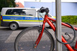 Ein rotes Fahrrad steht angeschlossen an einem Straßenschild, während im Hintergrund ein Polizeiauto parkt.