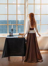 Red-haired Woman In Vintage Dress Stands Looks At Classic Window Waiting Love. Clothing Costume Countess Old Style White Blouse, Brown Long Skirt. Curly Red Hair. Redhead Girl Princess Back Rear View