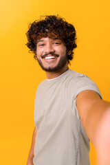 Wall Mural - Selfie time. Joyful carefree Indian young curly man talking selfie, looking at the camera and smiles, recording stories on his phone, vertical portrait