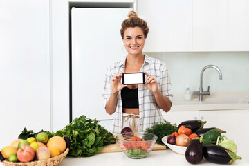 Woman is holding smartphone with blank screen for your design during cooking in modern kitchen