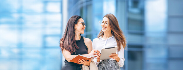 two young business lady with tablet and notebook walking outdoors near modern business center.