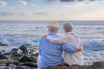 Canvas Print - Rear view of relaxed caucasian senior couple sitting on the pebble beach at sunset light admiring horizon over water. Two gray haired elderly people hugging with love