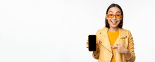 Portrait Of Stylish Korean Girl In Sunglasses, Smiling, Pointing Finger At Smartphone Screen, Showing Mobile Phone Application, Standing Over White Background