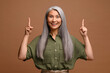 © Vadim Pastuh - Smiling mature grey-haired female business woman isolated on grey background points finger up. Lady paying attention at something