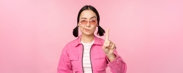 Image of serious, stylish asian girl in sunglasses, showing stop, prohibit gesture, taboo sign, saying no, standing over pink background