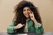 © SHOTPRIME STUDIO - Irritated stressed angry tanned curly Latin lady talking with phone sit at the table squeezes pen isolated over pastel beige background. Copy space Mockup Banner. Concept bad news problems