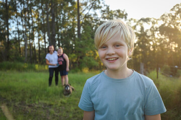 Wall Mural - Blonde caucasian boy smiling in the bush with mother and brother hugging out of focus in background