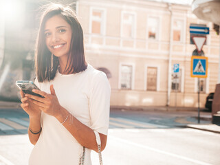 Wall Mural - Young beautiful smiling hipster woman in trendy summer dress. Sexy carefree woman posing on the street background at sunset. Positive model. Using smartphone apps. Looking at cellphone screen