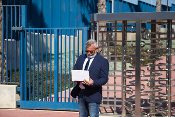 Mature male executive, grey-haired, bearded, with sunglasses, jacket and tie is consulting his laptop computer in the street. Concept businessman and executive.