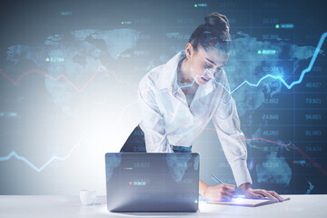 Wall Mural - Trading and investing concept with young businesswoman making notes in a book in front of laptop and digital screen with world map, indicators and raising financial chart graph. Double exposure