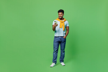Wall Mural - Full body smiling happy fun young man of African American ethnicity 20s in blue t-shirt hold in hand use mobile cell phone isolated on plain green background studio portrait. People lifestyle concept.