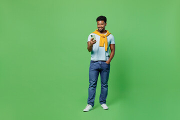 Full body smiling happy fun young man of African American ethnicity 20s in blue t-shirt hold in hand use mobile cell phone isolated on plain green background studio portrait. People lifestyle concept.