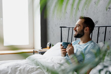 Side View Young Happy Minded Man Wear Blue T-shirt Lying In Bed Drink Coffee Tea Hold Cup Look Camera Rest Relax Spend Time In Bedroom Lounge Home In Own Room House Wake Up Dream. Real Estate Concept