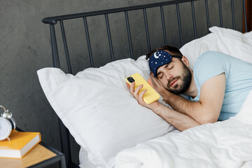 Wall Mural - Young man in blue t-shirt lying in bed close eyes sleep near mobile cell phone hands folded under head rest relax spend time in bedroom lounge home in own room house wake up dream Real estate concept