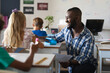 © wavebreak3 - Smiling african american young male teacher talking to caucasian elementary schoolgirl at desk