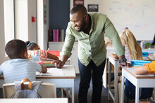 Smiling African American Young Male Teacher Talking To African American Elementary Boy At Desk