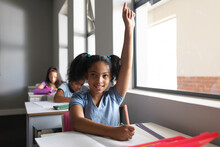 Smiling Biracial Elementary School Girl With Hand Raised Sitting At Desk In Classroom