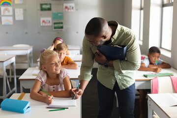 Wall Mural - Smiling african american young male teacher assisting caucasian elementary schoolgirl at desk