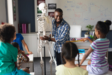 Wall Mural - Happy african american young male teacher explaining skeleton to multiracial elementary students