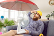 Leinwandbild Motiv Funny sweaty chubby bearded man in swimsuit sitting at home, suffering from crazy summer heat, wiping sweat off forehead, holding electric fan, wishing for heatwave to stop and fresh breeze to blow