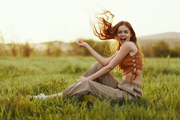 A woman sits on the green young fresh grass in the open air and laughs and smiles happily in the sunset light, her flying red hair fluttering in the wind