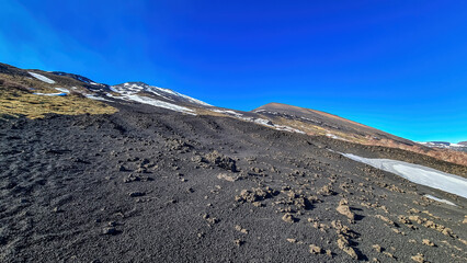 Solidified lava, ash and pumice on its slopes of snow covered craters. Yellow golden dry grass on brown dark volcanic sand, bare terrain. Scenic view on volcano mount Etna, in Sicily, Italy, Europe.