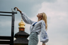 Young Woman Stands In Ukrainian Embroidered Shirt Against Background Of Church Dome And Sky.
