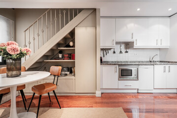 Sticker - Long apartment kitchen with white cabinets, pink granite countertops, built-in appliances, brown hardwood parquet floors, round dining table and stairs up to bedrooms