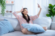 © Basicdog - Smiling Asian woman happily sits in the living room and enjoys taking selfies.