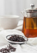 Strainer infusor with loose black tea on light cloth background with clear glass teapot and white ceramic cup