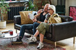 © AnnaStills - Modern mature Caucasian couple sitting on couch in loft living room taking to friends or relatives on video call using digital tablet
