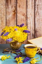 Bouquet Of Yellow Primroses And Muscari In A Vase. Wooden Background, Book, Yellow Cup, Glasses. Blue Tablecloth. Side View.
