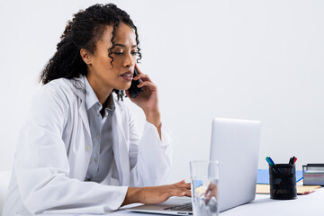 Wall Mural - African american mid adult woman talking over smart phone and using laptop on desk, copy space