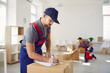 © Studio Romantic - Professional male mover in overalls makes notes on clipboard while standing near cardboard boxes. Worker of relocation service makes consignment note against background of colleague who packs boxes.