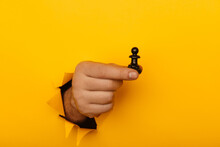 Hand Is Holding Figure Of Black Pawn Through A Hole Of Yellow Background
