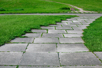 Wall Mural - park path made of square tiles overgrown with grass in a parkland with a green lawn close up of the way on park with meadow and asphalt road lit by sun, nobody.