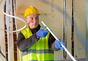 Wall Mural - Caucasian electrician in vest and helmet standing with cable duct in hands and looking through it.