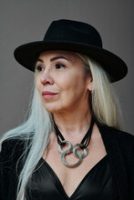 Vertical Medium Close-up Studio Portrait Of Beautiful Mature Woman With Long Gray Hair Wearing Modern Goth Outfit With Hat Looking Away
