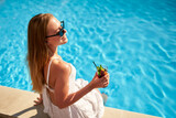 Fototapeta  - Blonde woman drinks refreshing cocktail sunbathing and sitting near swimming pool at tropical spa. Female in sunglasses enjoys a drink on poolside. Girl chilling in tropical resort on vacation.