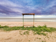 Empty Bench By The Sea On A Cloudy Day. View Of A Bench On An Empty Sandy Beach On The Shore Of The Sea Of Azov Under A Cloudy Sky On A Cool Day.