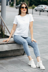 Wall Mural - Stylish brunette girl wearing white t-shirt, blue jeans and posing sitting on a bench , urban clothing style. Street photography